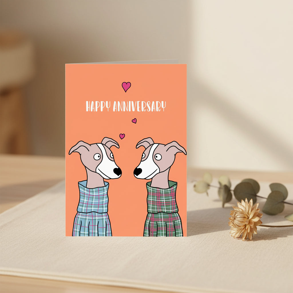 A greeting card with a bright coral-orange front featuring the words “HAPPY ANNIVERSARY” above two illustrated dogs facing each other, with small pink hearts between them. The card is shown with a bright pink envelope or backing behind it on a white background.
