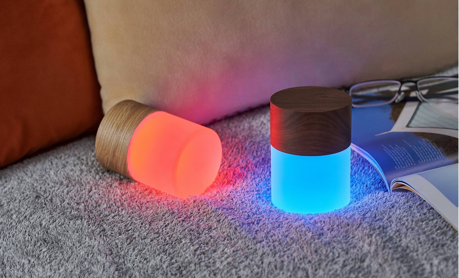 Two Lemelia lights on grey carpet beside a book, glowing coral and blue