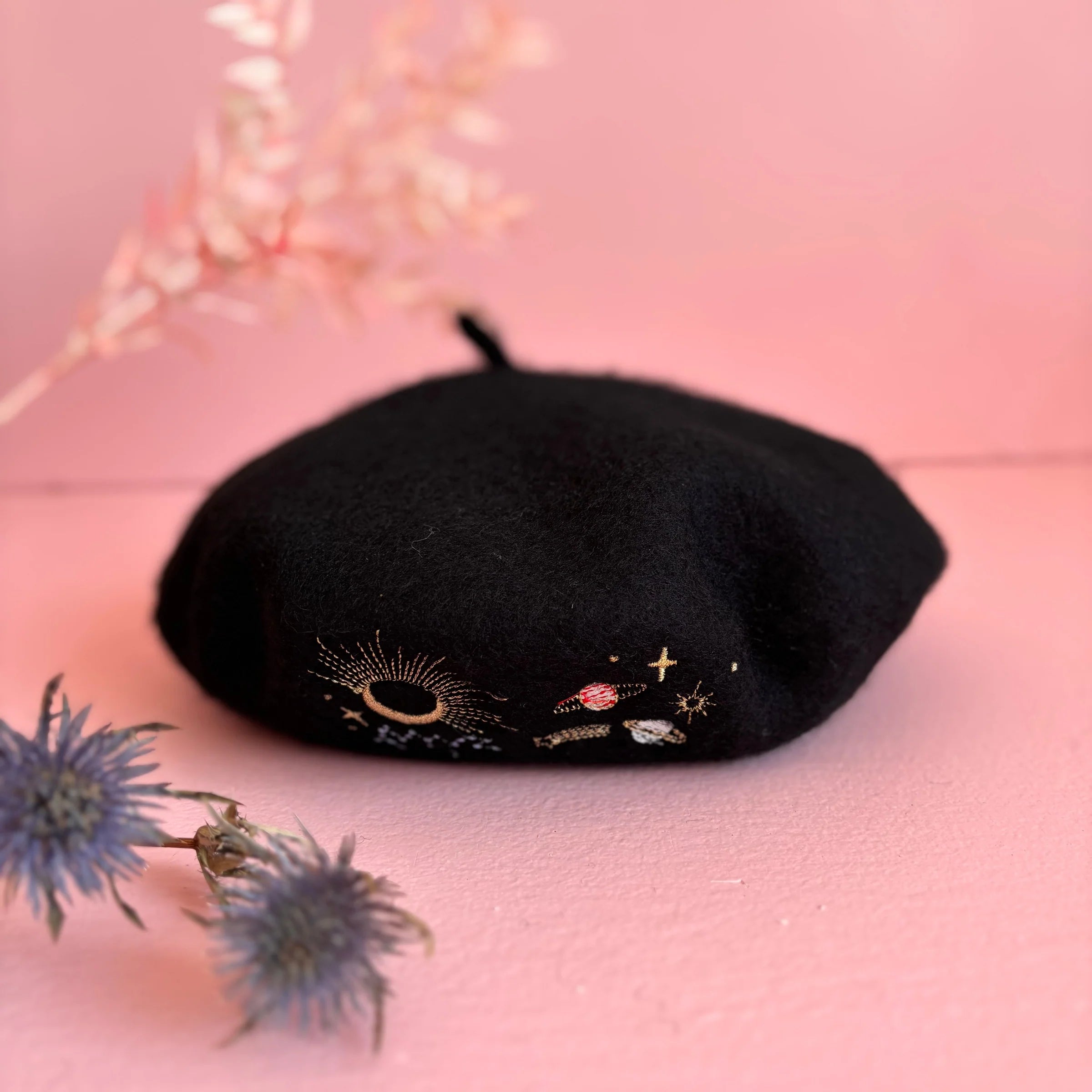 Black beret with embroidered galaxy design displayed on pink background.