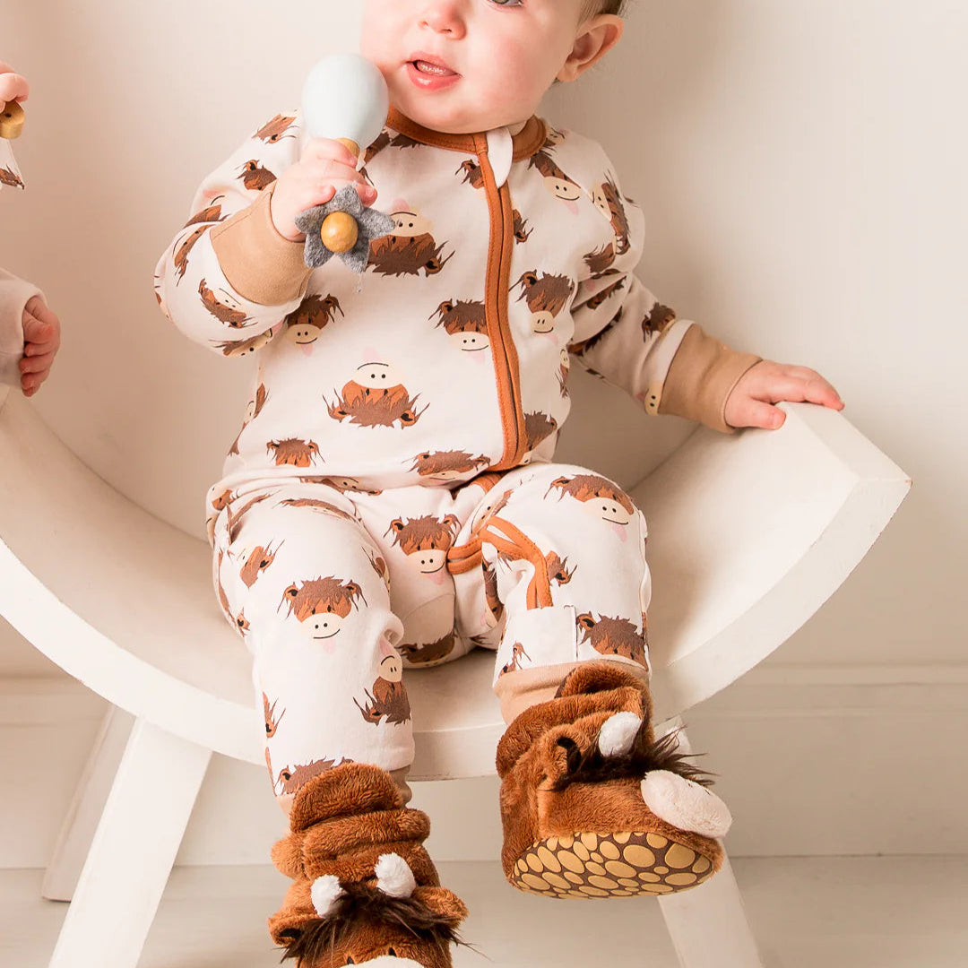 Baby sitting in a chair wearing the Neutral Hamish Highland Cow romper with matching cow slippers, looking up and smiling.
