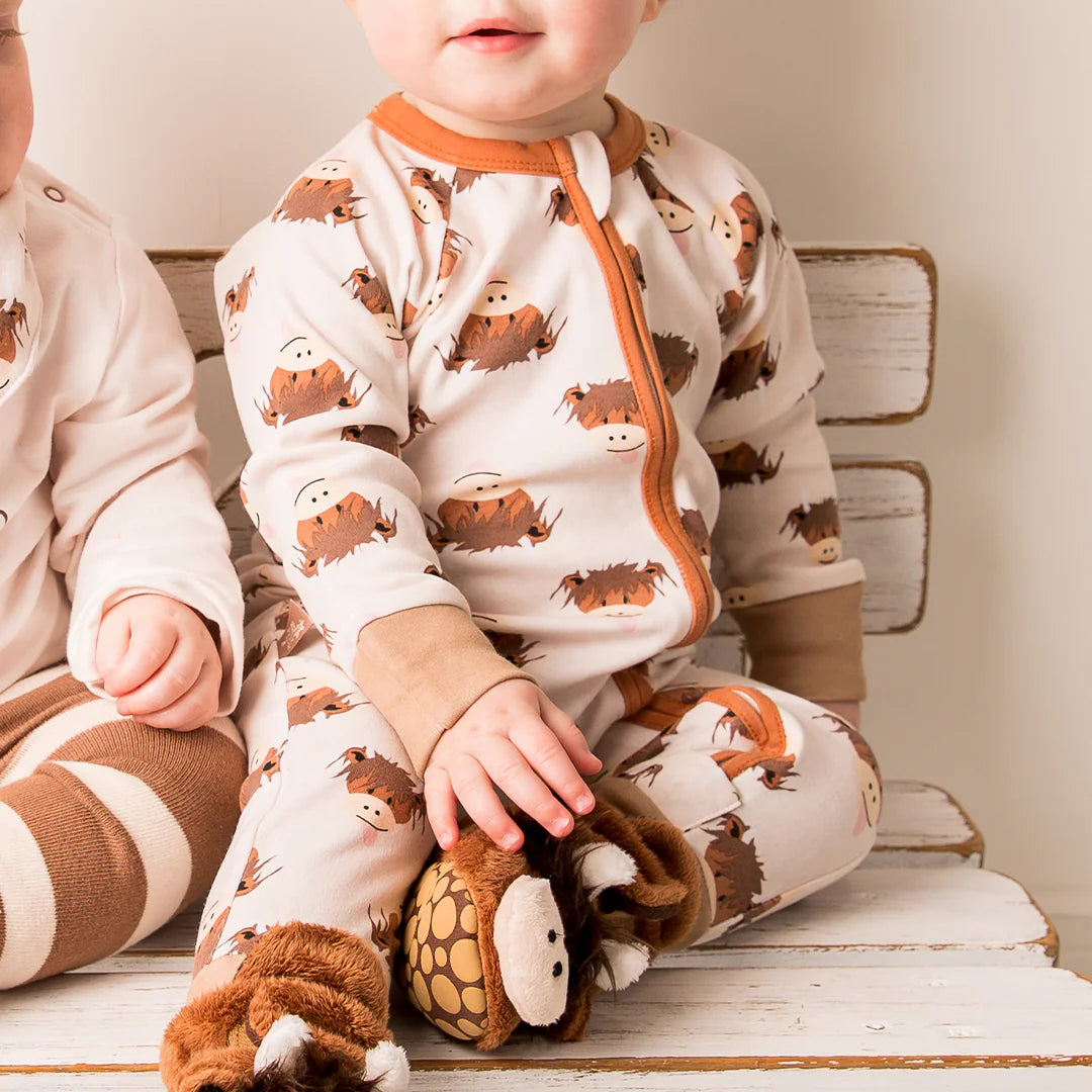 Baby sitting on a bench in the Neutral Hamish Highland Cow zip-up romper and slippers, facing forward with a gentle smile.