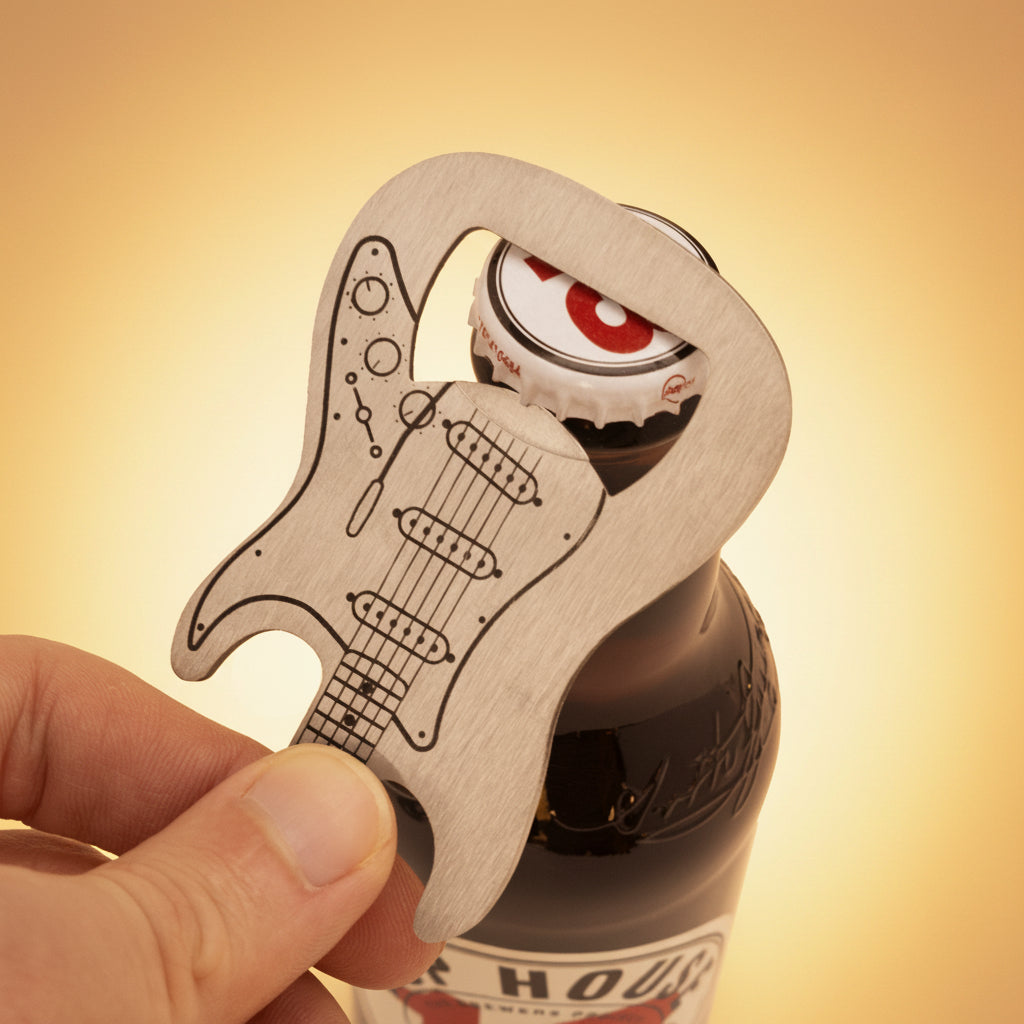 Close-up of hand using stainless steel electric guitar bottle opener to remove the cap from a brown glass beer bottle.