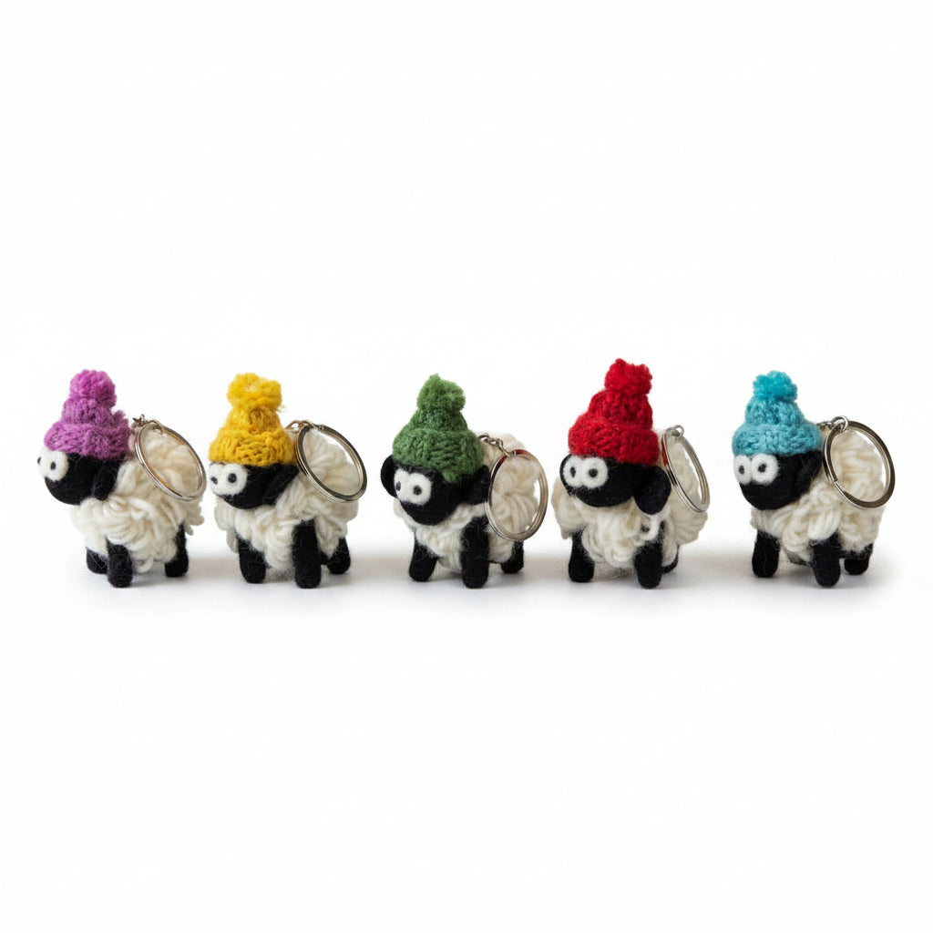 Five small sheep-shaped keychains lined up on a gray slate board against a light gray background. Each sheep has a black face and body, white wool, and a different colored knit hat in purple, yellow, green, red, and blue.