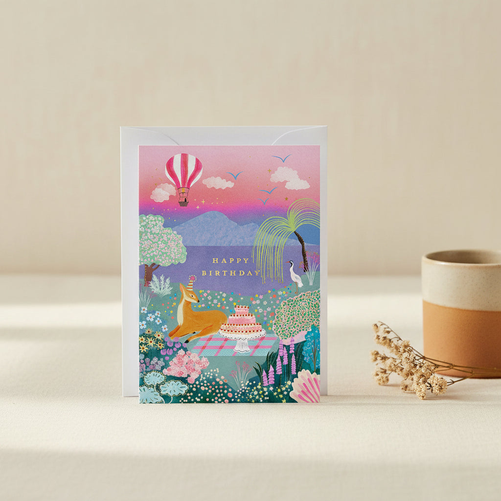 Birthday card with a deer, cake, and hot air balloon in a colorful landscape