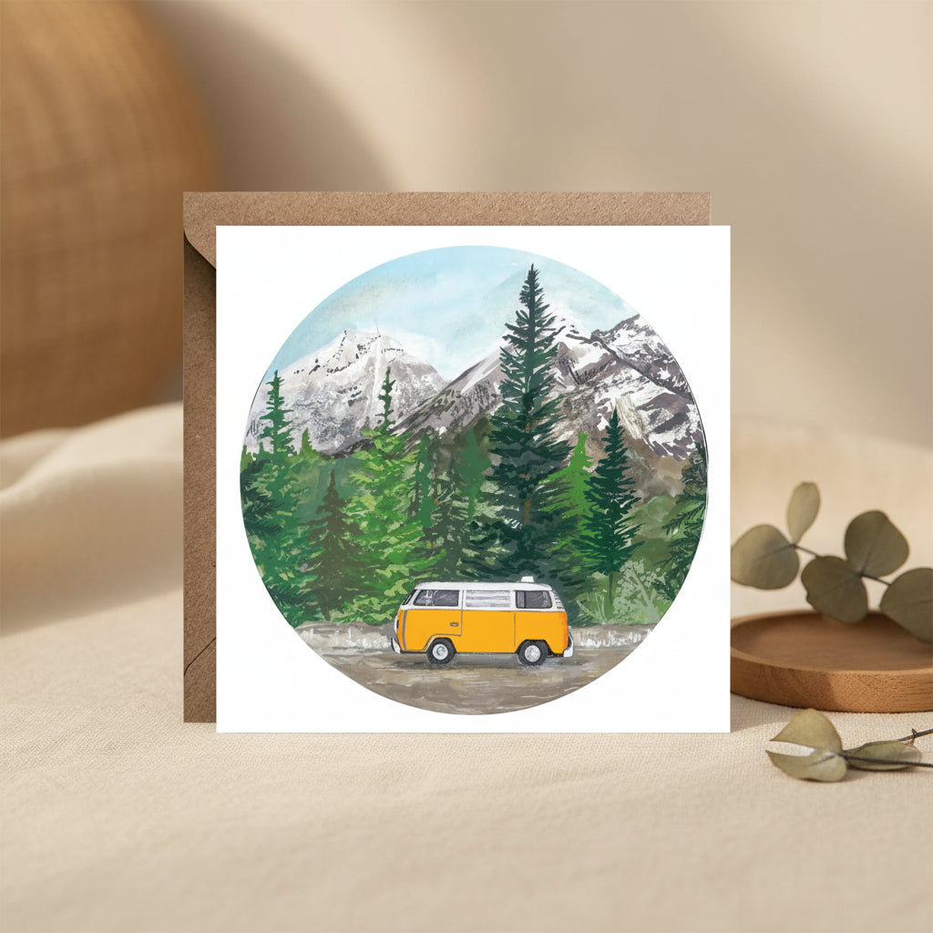A square greeting card with a kraft envelope, featuring a circular watercolor illustration of a yellow vintage camper van parked in front of a snowy mountain and pine forest.