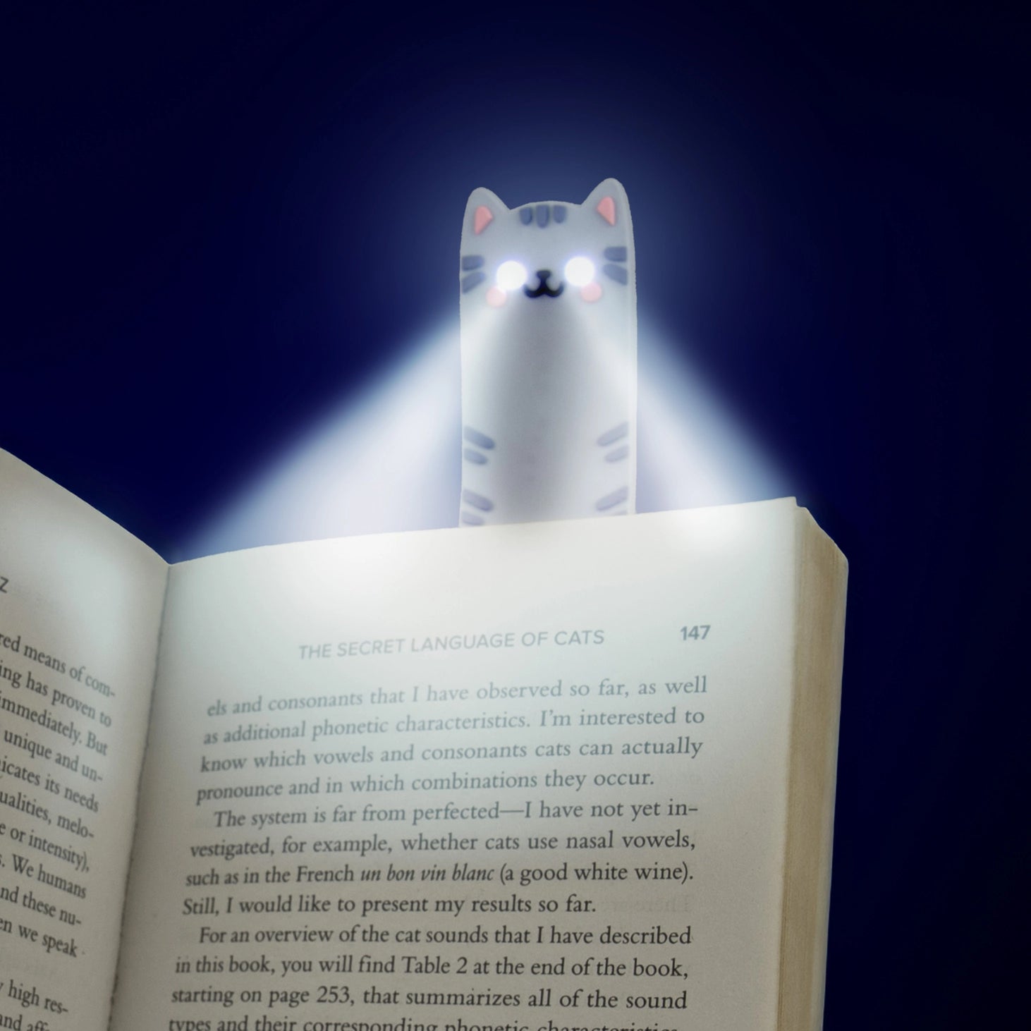 Cat reading light glowing with bright white LED beams in a dark room, clipped to the top of an open book for nighttime reading.