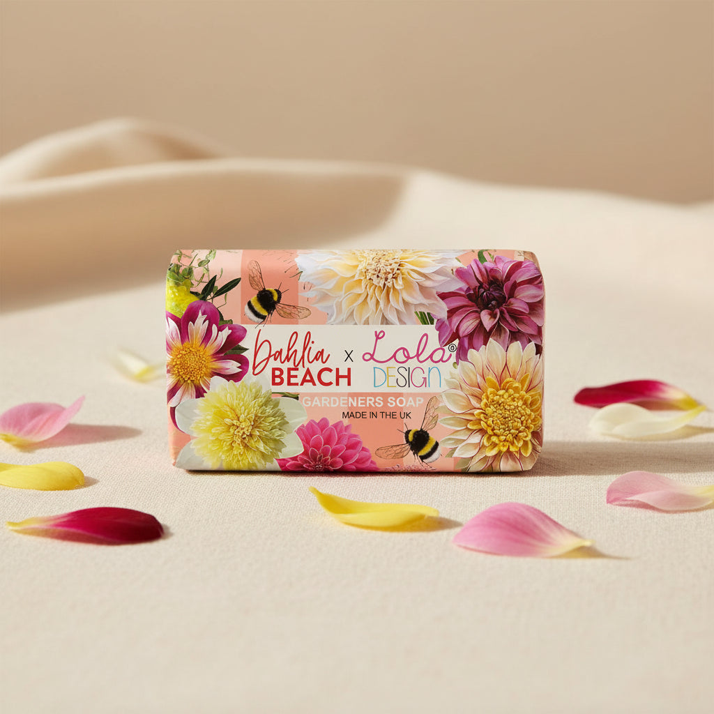 A rectangular bar of Dahlia Beach x Lola Design Gardeners Soap on a white background. The soap features a vibrant floral pattern with pink, yellow, and white dahlias, bumblebees, and a central white label with red and blue text.