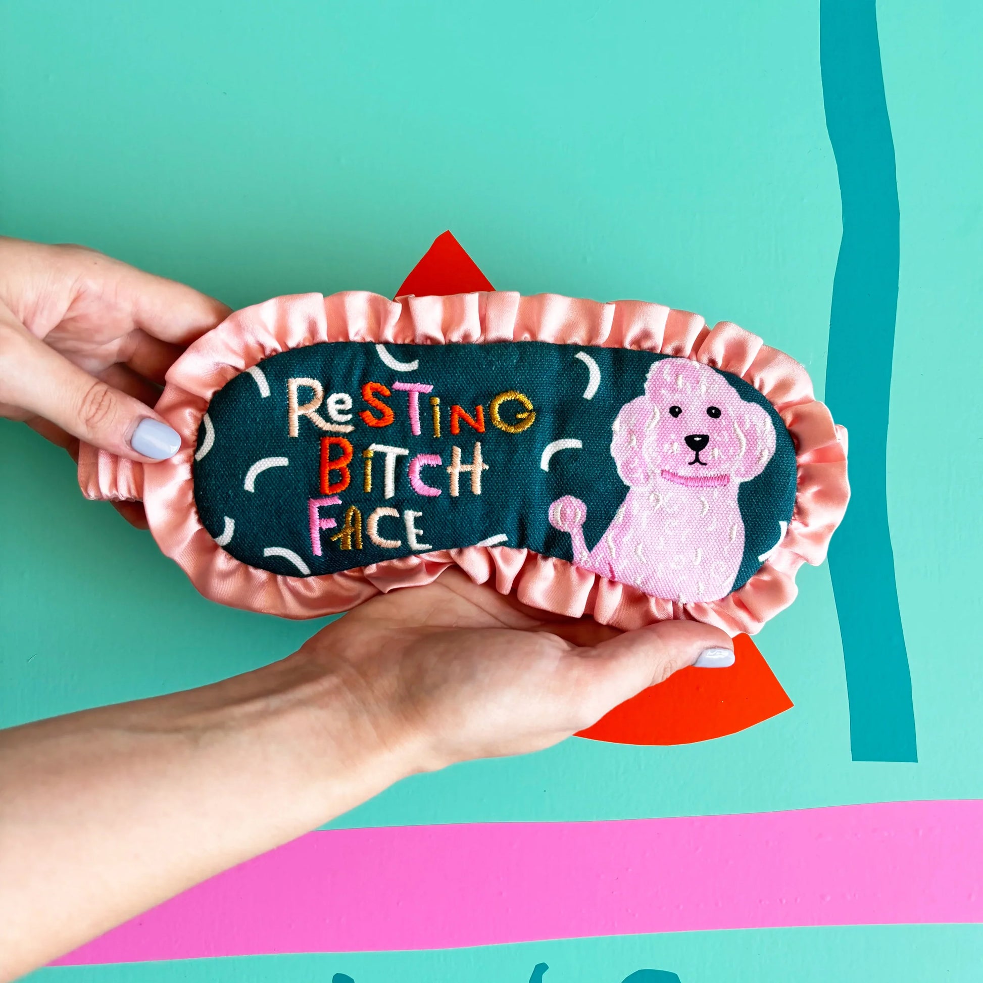 Resting Bitch Face eyemask with pink satin trim and embroidered dog design held in hands.
