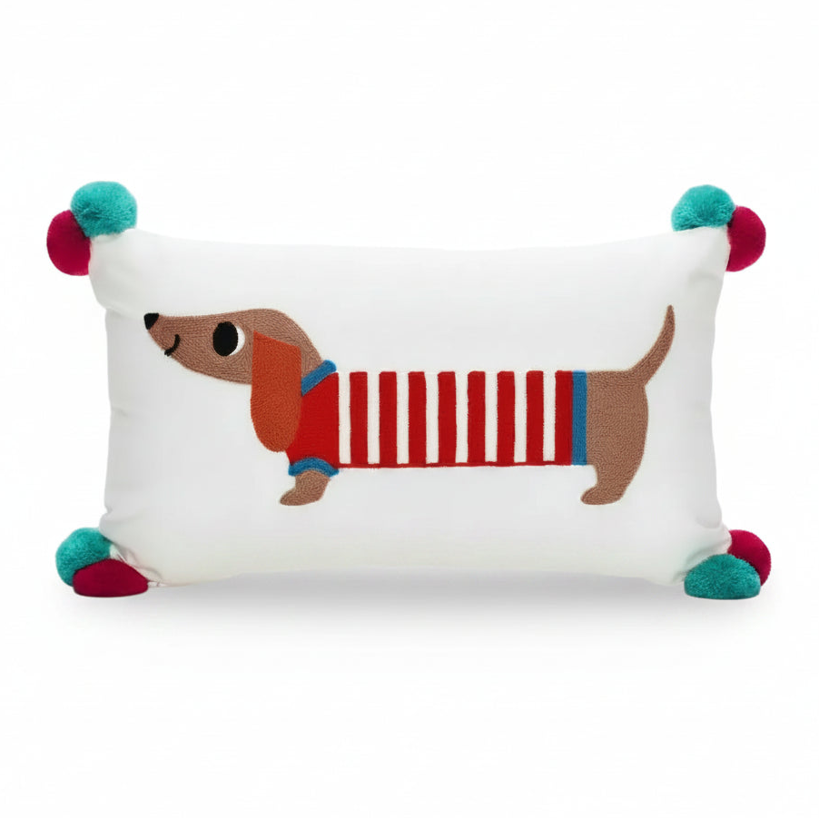 White oblong embroidered cushion with sausage dog design and red and blue pom poms on corners.