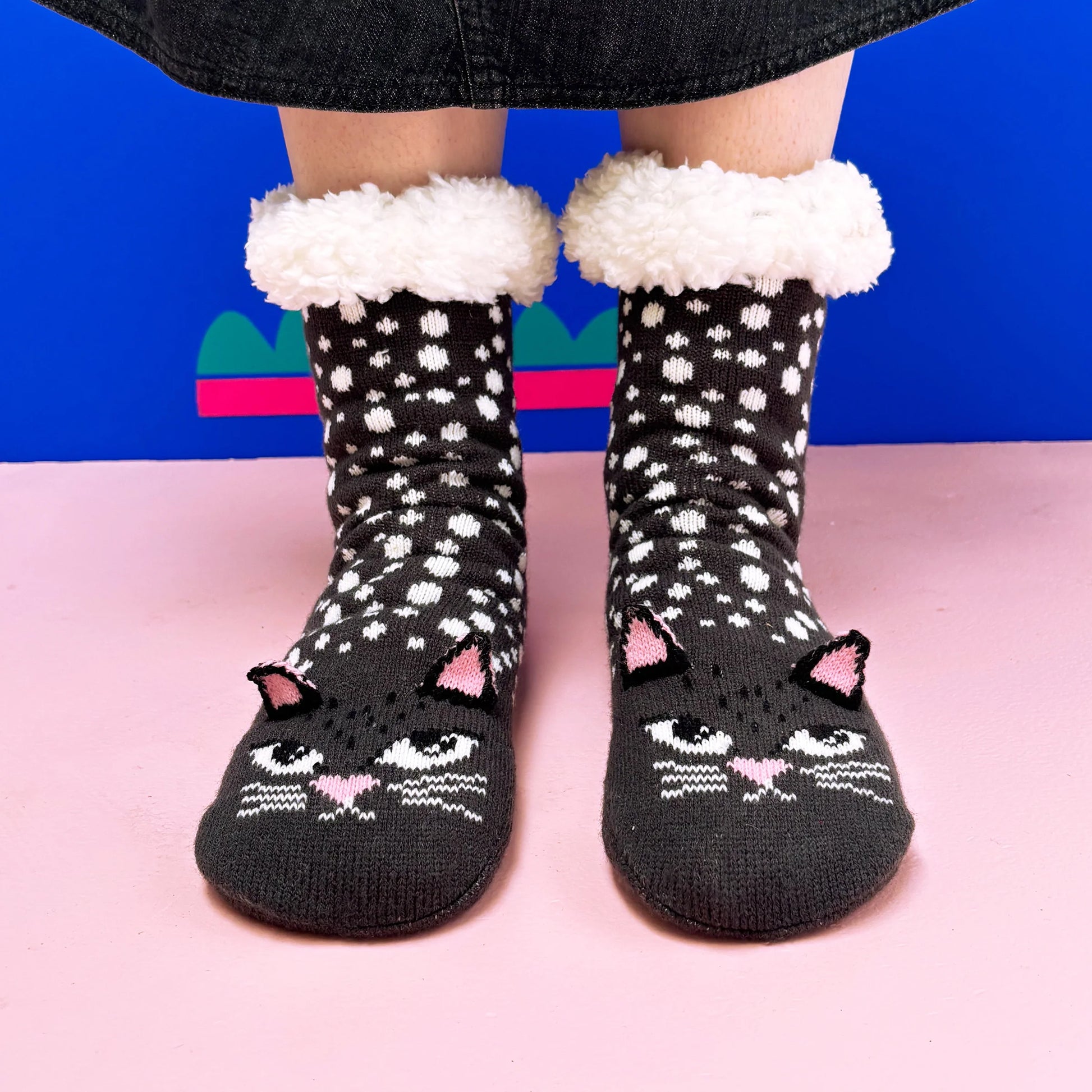 Model wearing black cat face slipper socks with fluffy cuffs on pink floor