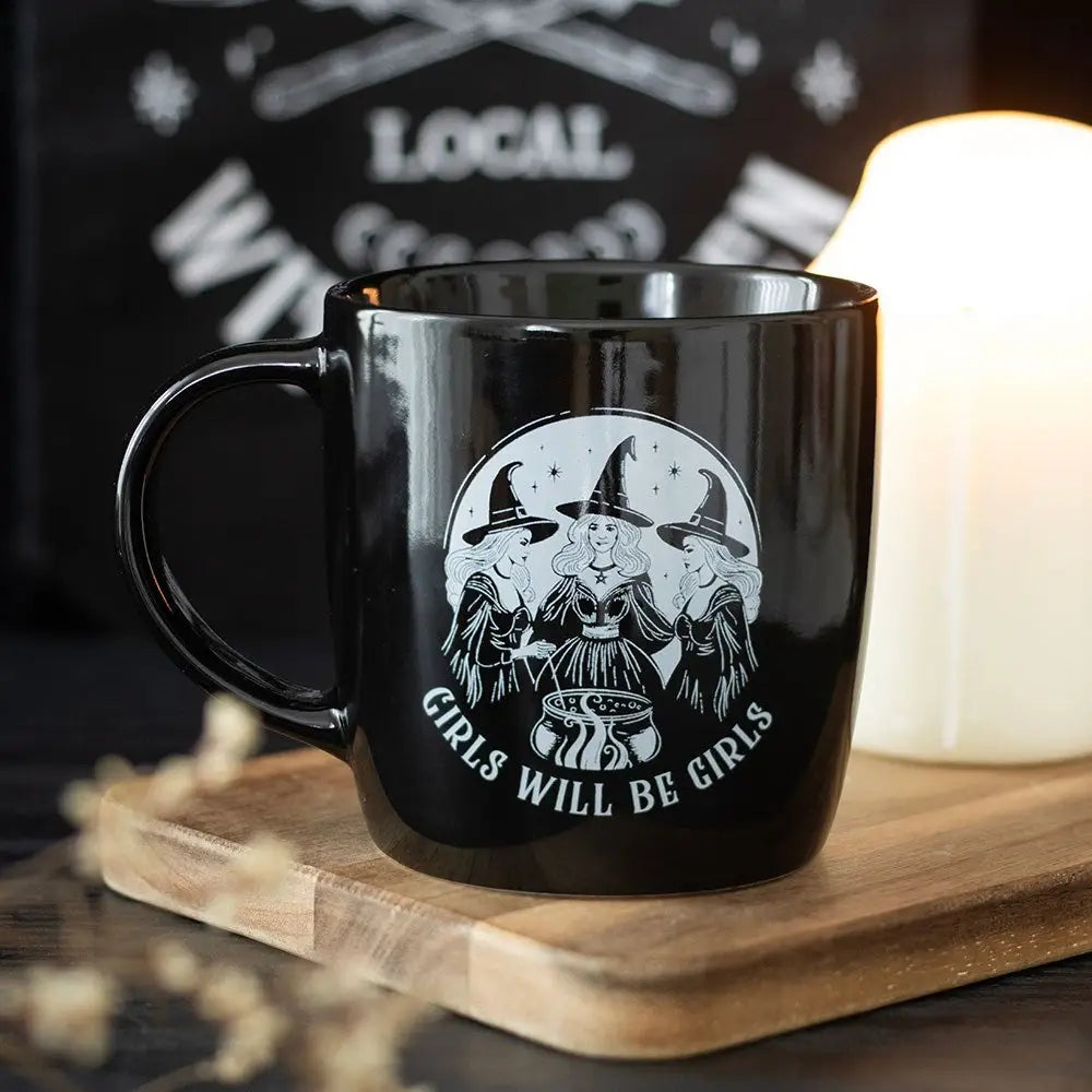 Black ceramic mug with ‘Girls Will Be Girls’ witch design on wooden board beside candle.