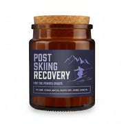 An amber glass jar candle with a cork lid and a dark navy label reading 'POST SKIING RECOVERY' with a skier graphic, set against a snowy mountain background.