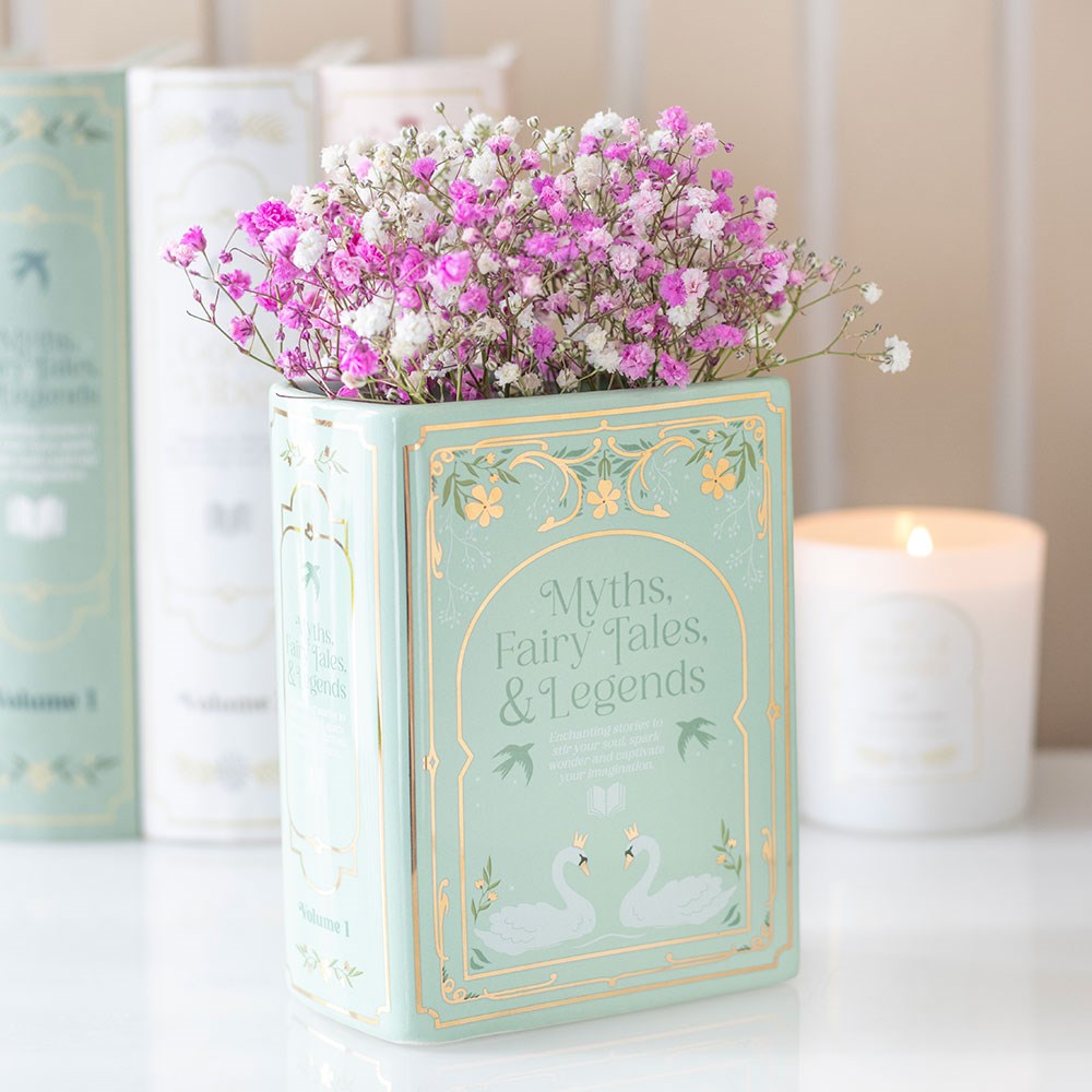 Green ceramic book shaped vase with pink and white flowers displayed on a shelf beside books and a lit candle – literary home décor for book lovers.