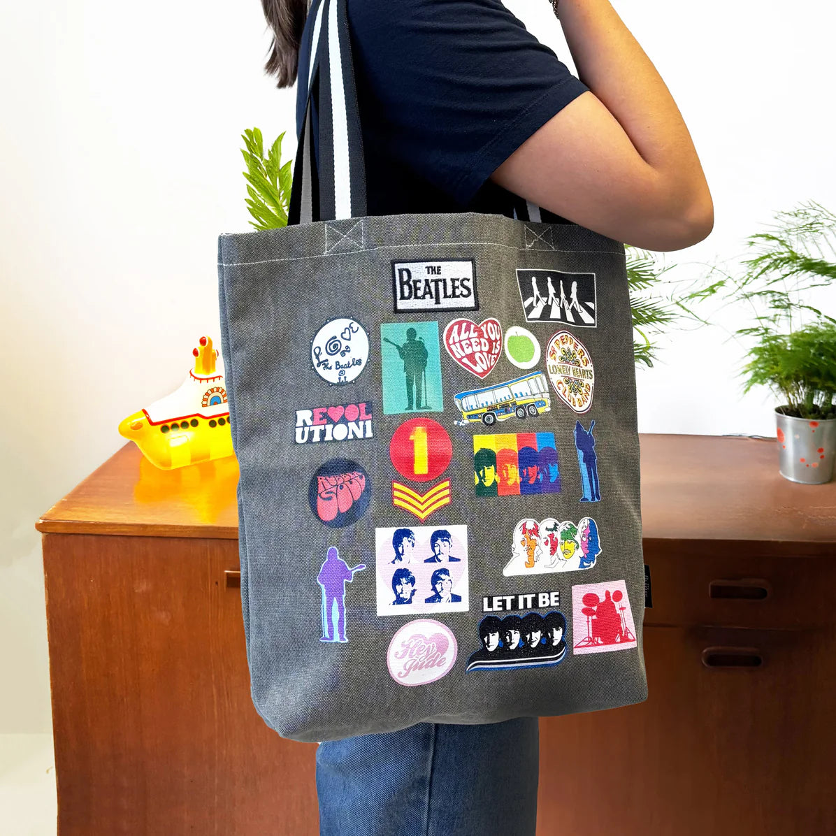 Person wearing grey Beatles tote bag with colourful printed patch designs and black-and-white straps, next to a yellow submarine model.