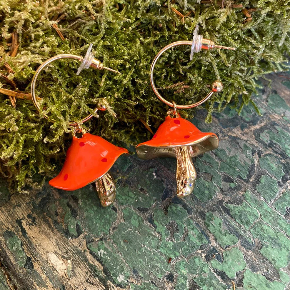 Pair of Forage Red Earrings resting on moss and textured green surface, showing curved golden stems and bright red mushroom caps with dotted detail.