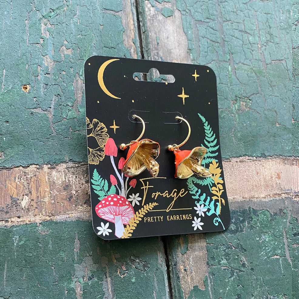 Angled view of Forage Red Earrings on branded card, highlighting golden mushroom gills and red enamel caps against forest-themed packaging.