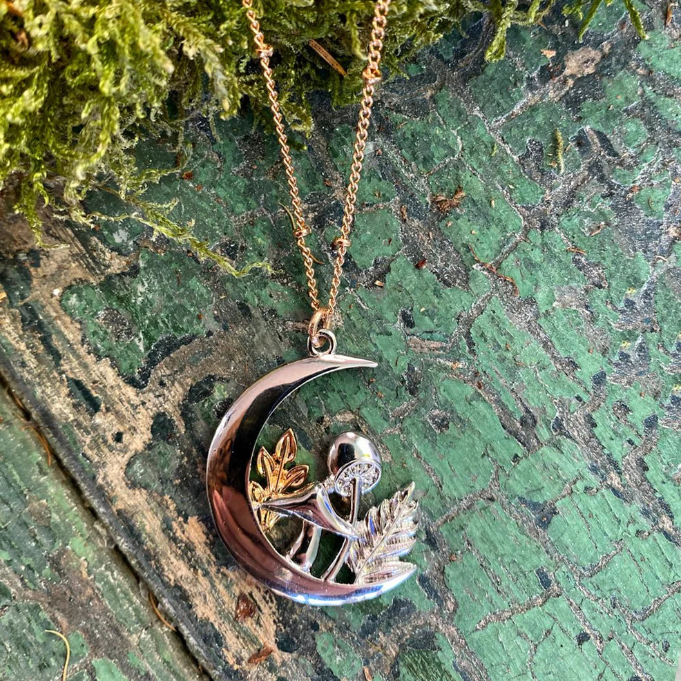  Forage Moon Necklace laid on distressed green wood with moss, featuring a silver crescent moon pendant with gold and silver mushroom and leaf details on a gold chain.