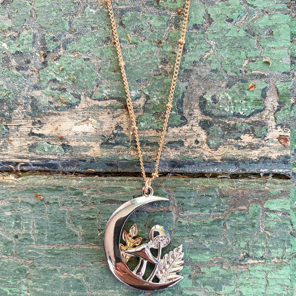 Close-up of Forage Moon Necklace pendant on rustic green background, highlighting the intricate mushroom and leaf detailing inside the silver crescent moon.