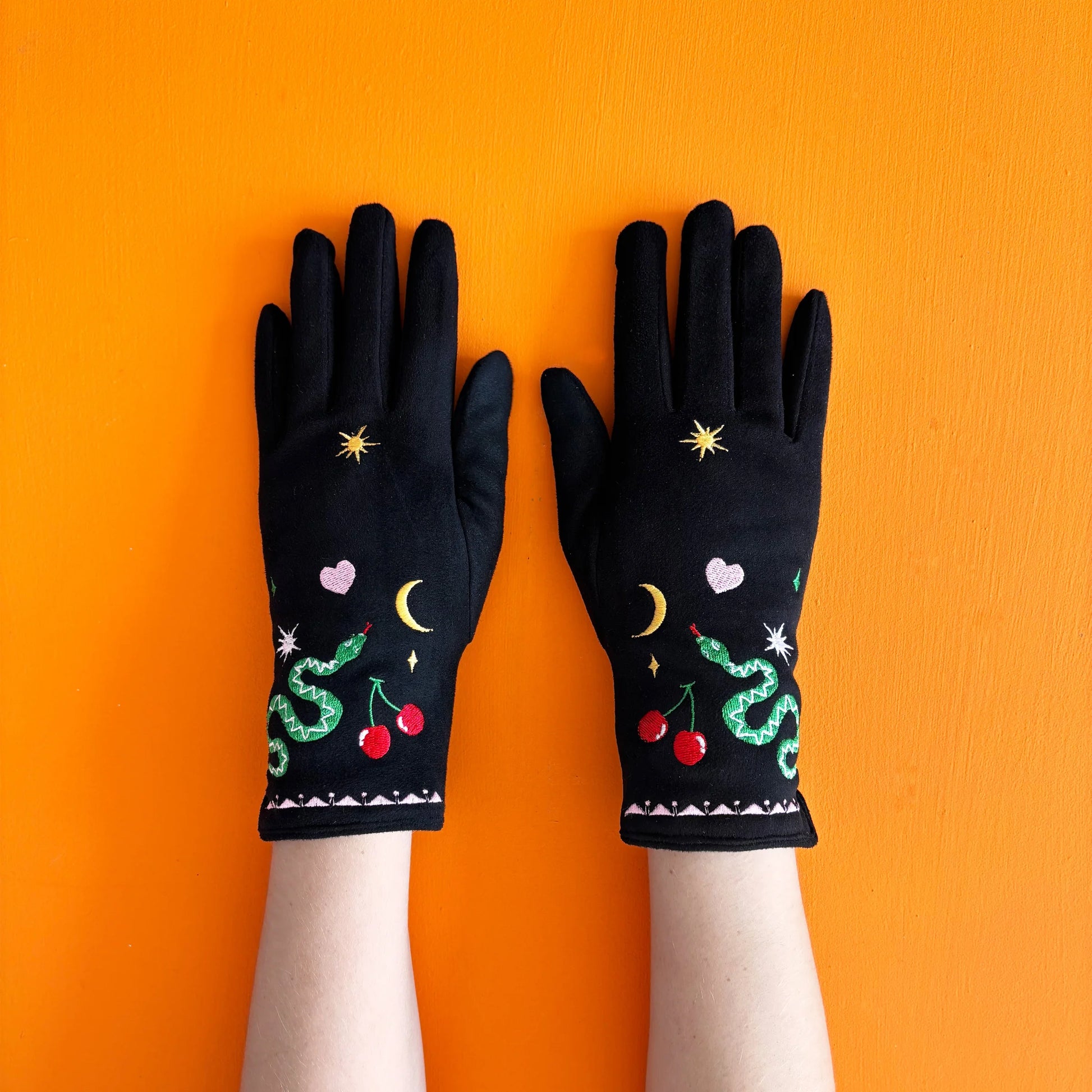 Black suedette embroidered gloves with snake, cherry, and heart designs, displayed flat on a bright orange background.
