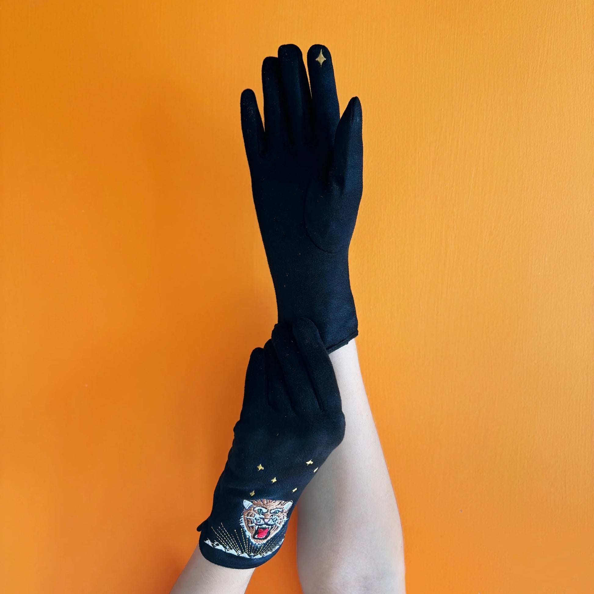 Model wearing black suedette tiger gloves with gold embroidery and star on fingertip, arms crossed against orange background.