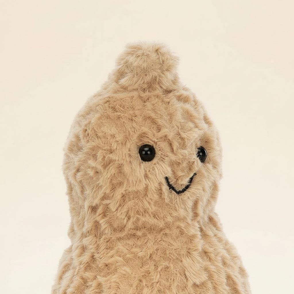 A plush peanut-shaped stuffed animal in a light tan/beige color with a brown corduroy-look foot and a simple embroidered smiley face, photographed on a clean white background.