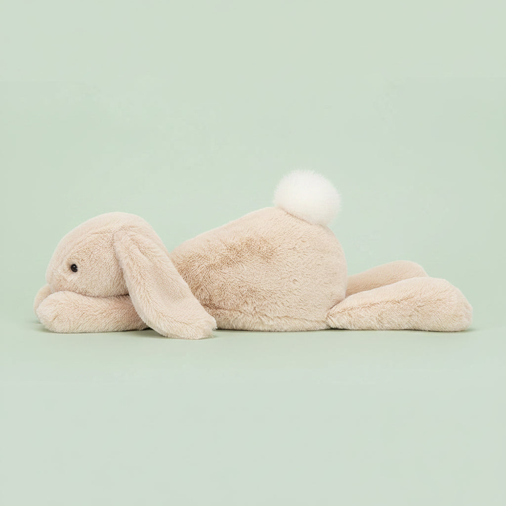 A side view of Jellycat Smudge Rabbit with oatmeal coloured fur, long floppy ears and a white fluffy tail, lying stretched out.
