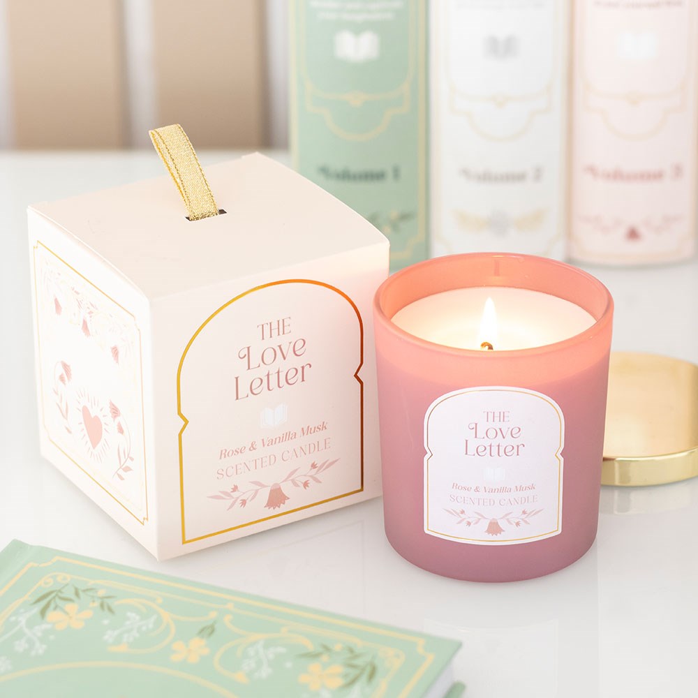 The Love Letter Rose & Vanilla Musk Candle lit in pink glass jar beside illustrated gift box with gold lid and romantic book-style packaging.