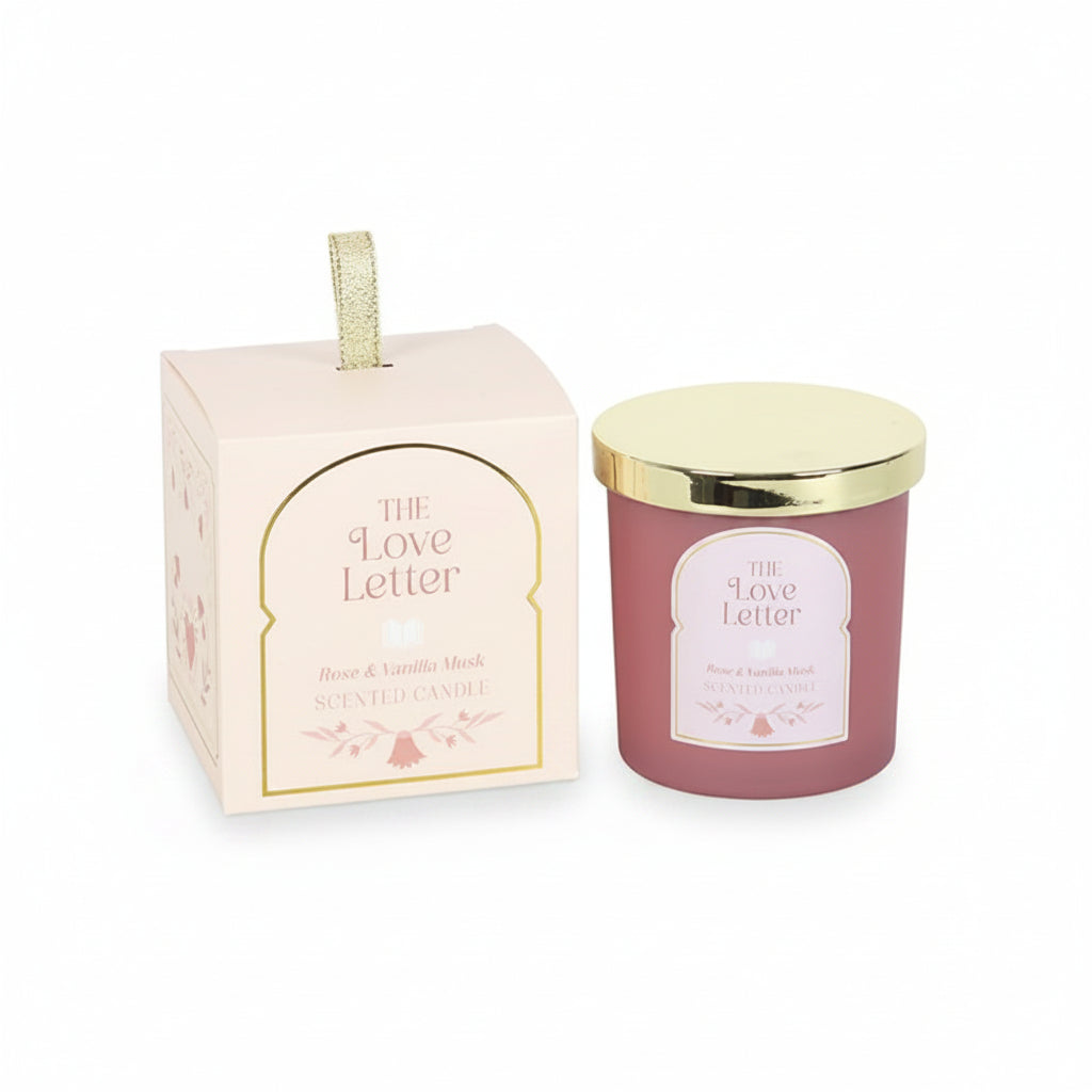The Love Letter Rose & Vanilla Musk Candle in pink jar with gold lid displayed next to decorative illustrated gift box with gold ribbon handle.