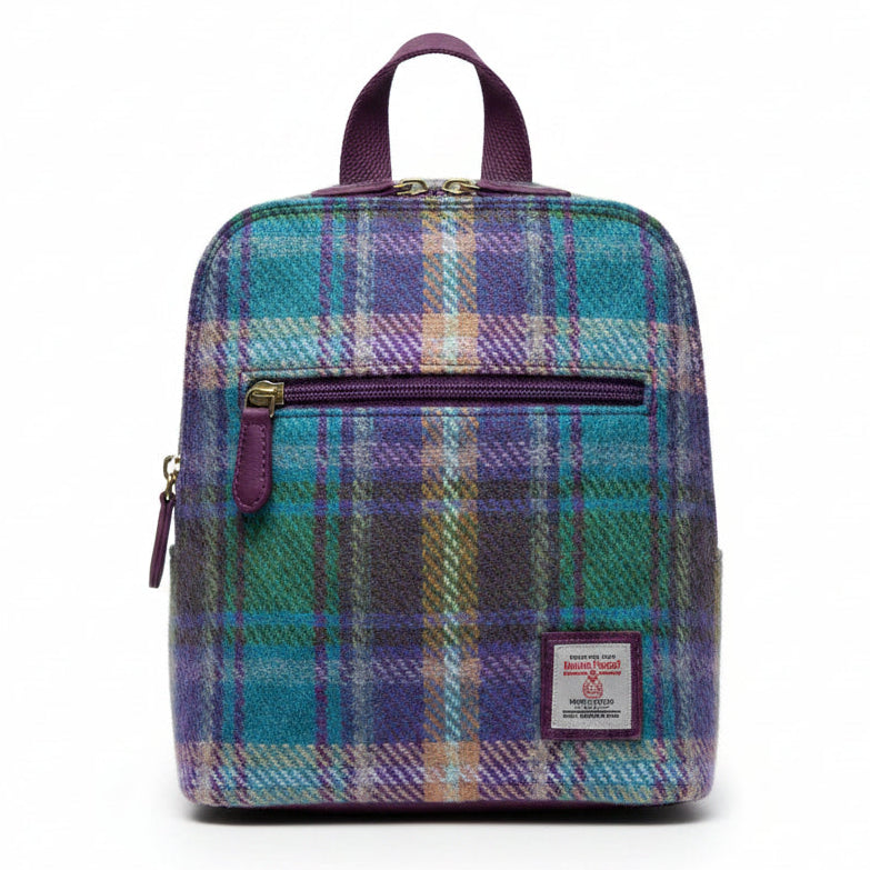 A person wearing a chunky gray knit sweater carries a small plaid backpack in a sunny outdoor setting. The backpack features a multicolor tartan pattern with shades of blue, purple, and green, and has a visible 'The Design Museum' label on the front.