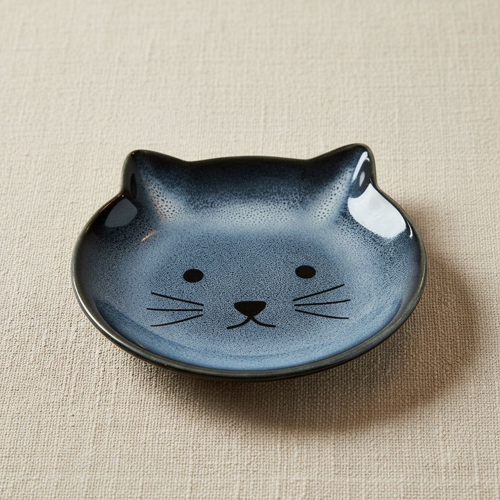 A small round ceramic dish shaped like a cat's face, featuring a speckled dark gray and black glaze with simple black facial details, shown on a plain white background.