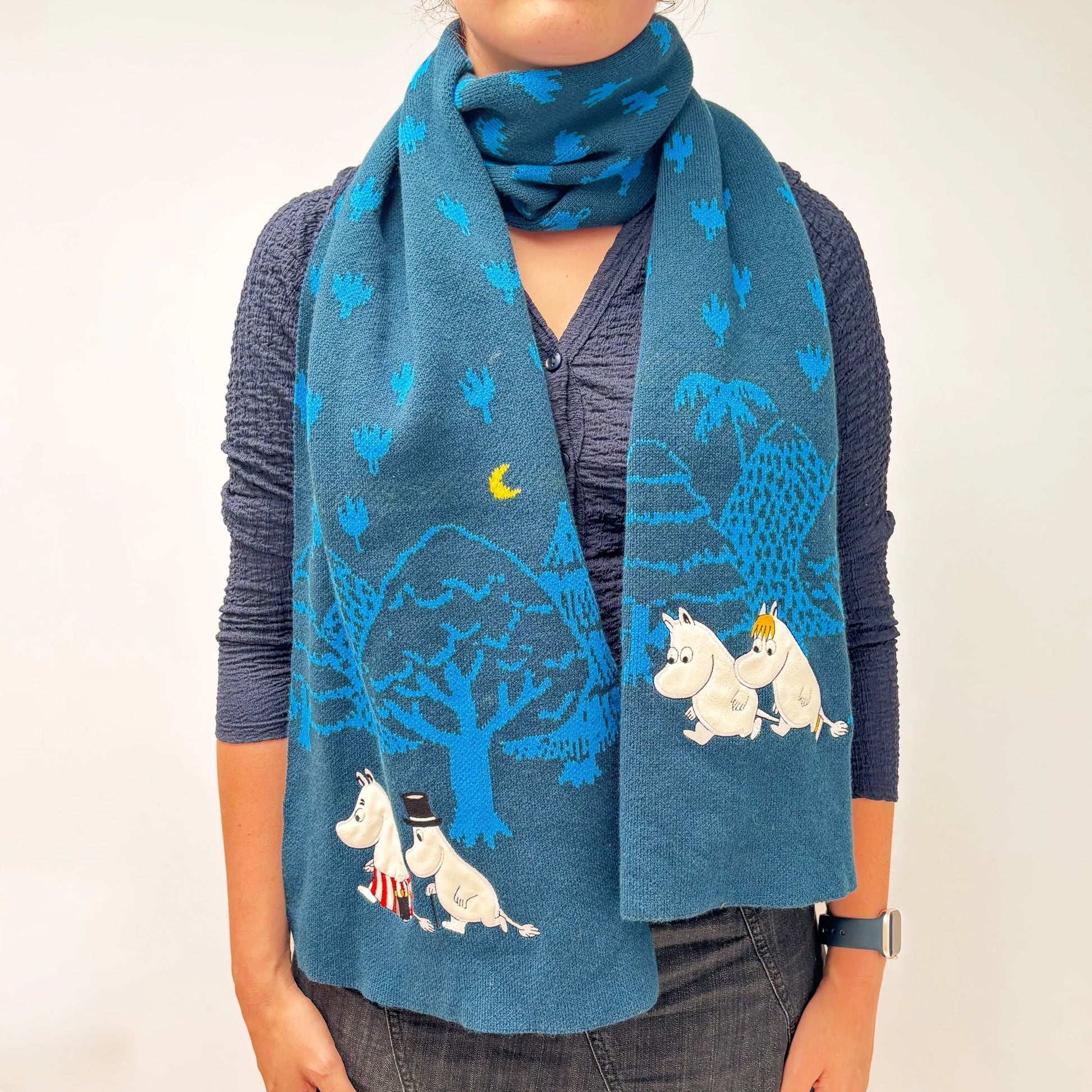 Person wearing Moomin Blue Forest Embroidered Scarf with forest and moon design in soft blue tones.