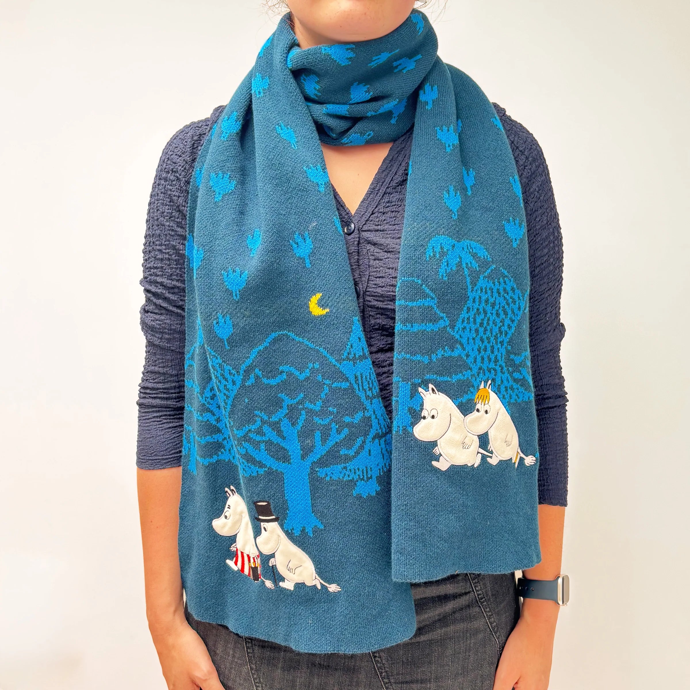 Person wearing Moomin Blue Forest Embroidered Scarf with forest and moon design in soft blue tones.