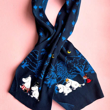 Knitted Moomin scarf in blue with embroidered characters and forest design, displayed against pink background.