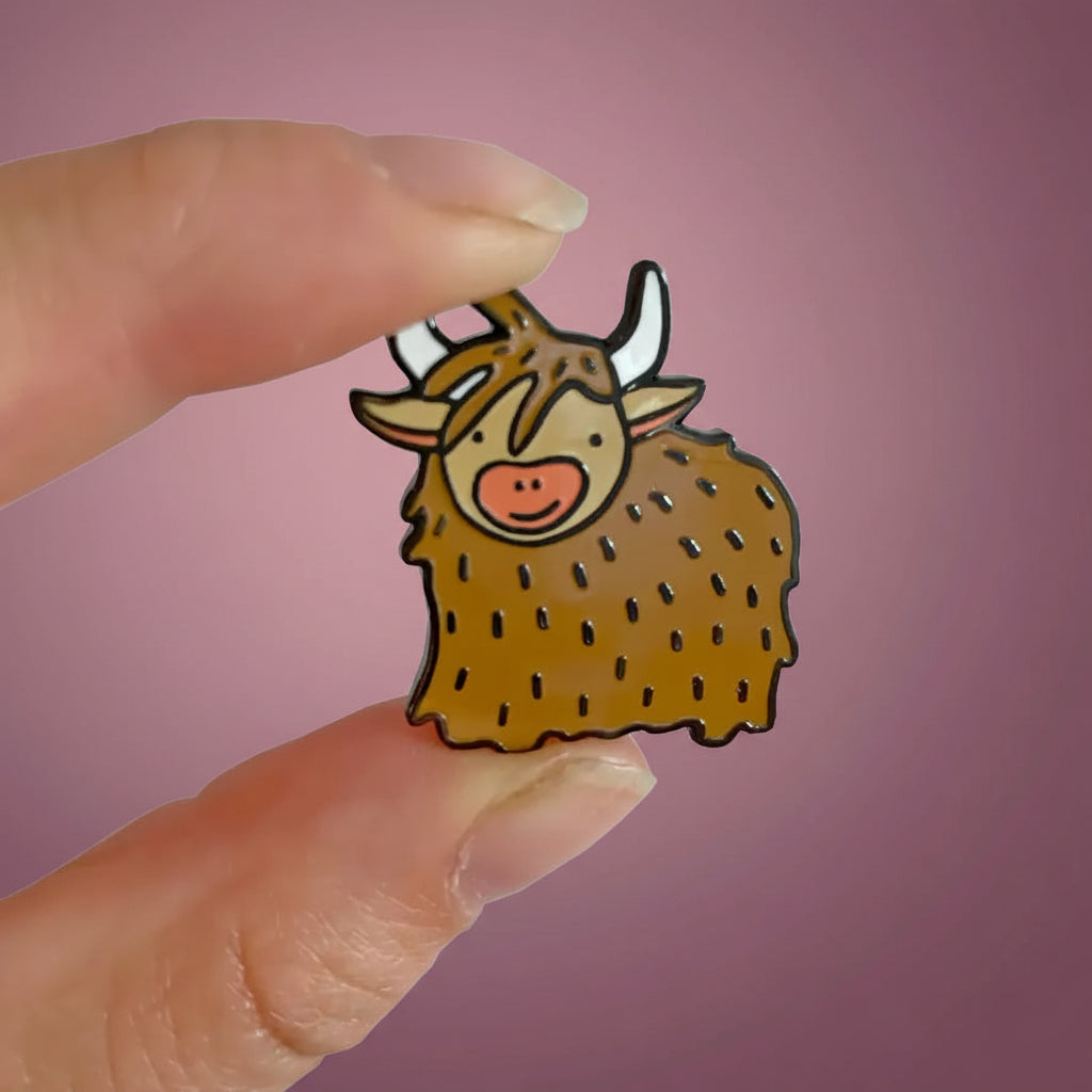 Cute enamel Highland Cow pin badge with cartoon-style design, shown being held between two fingers for scale – Scottish accessory for bags, hats, and clothes.