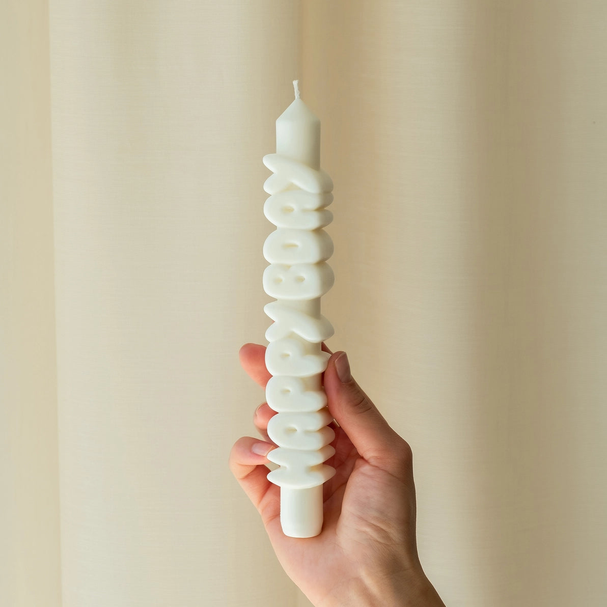 Person holding a white 9-inch handmade taper candle with raised 3D “HAPPY BDAY” lettering, made from soy-paraffin wax blend, against a neutral curtain background.