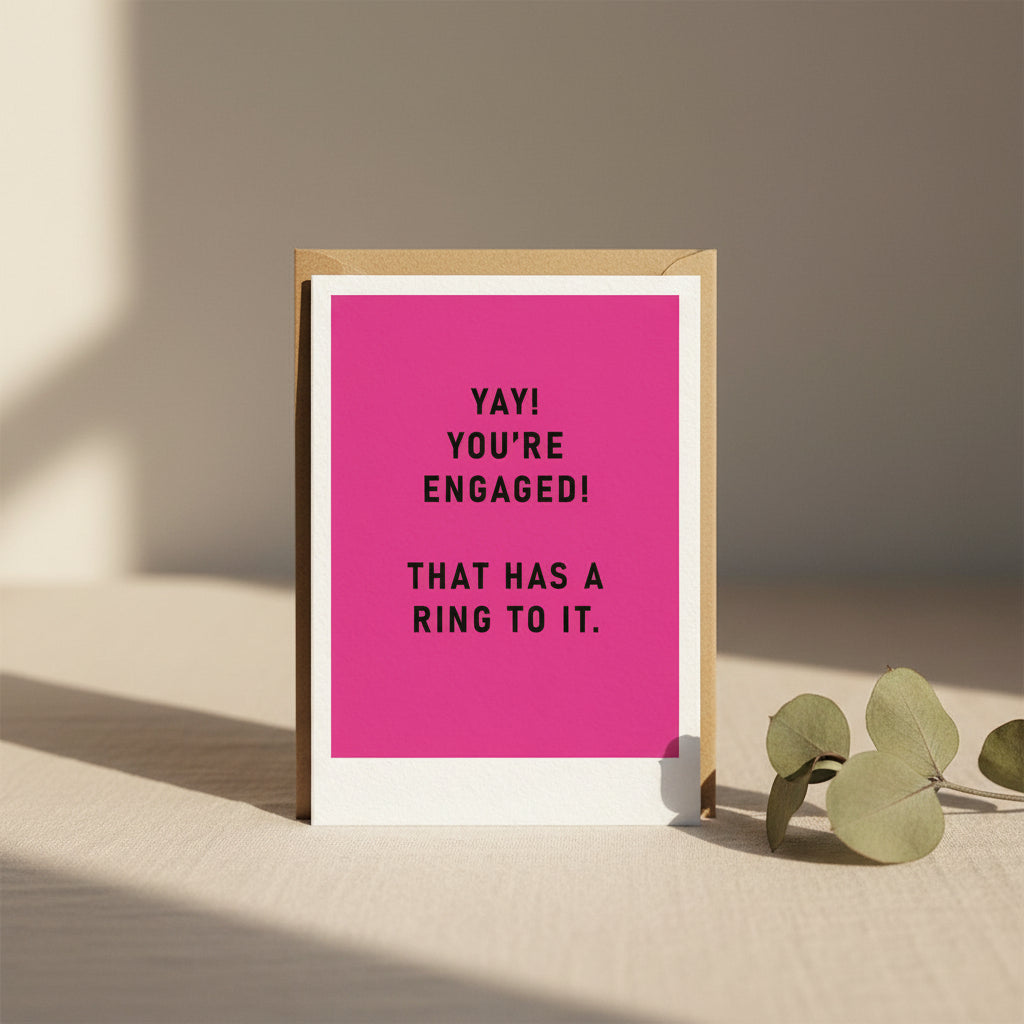 Pink card with black text 'YAY! YOU'RE ENGAGED! THAT HAS A RING TO IT.' on a white background