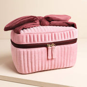 Pale pink velvet makeup bag with burgundy bow handle and gold zip on cream background.