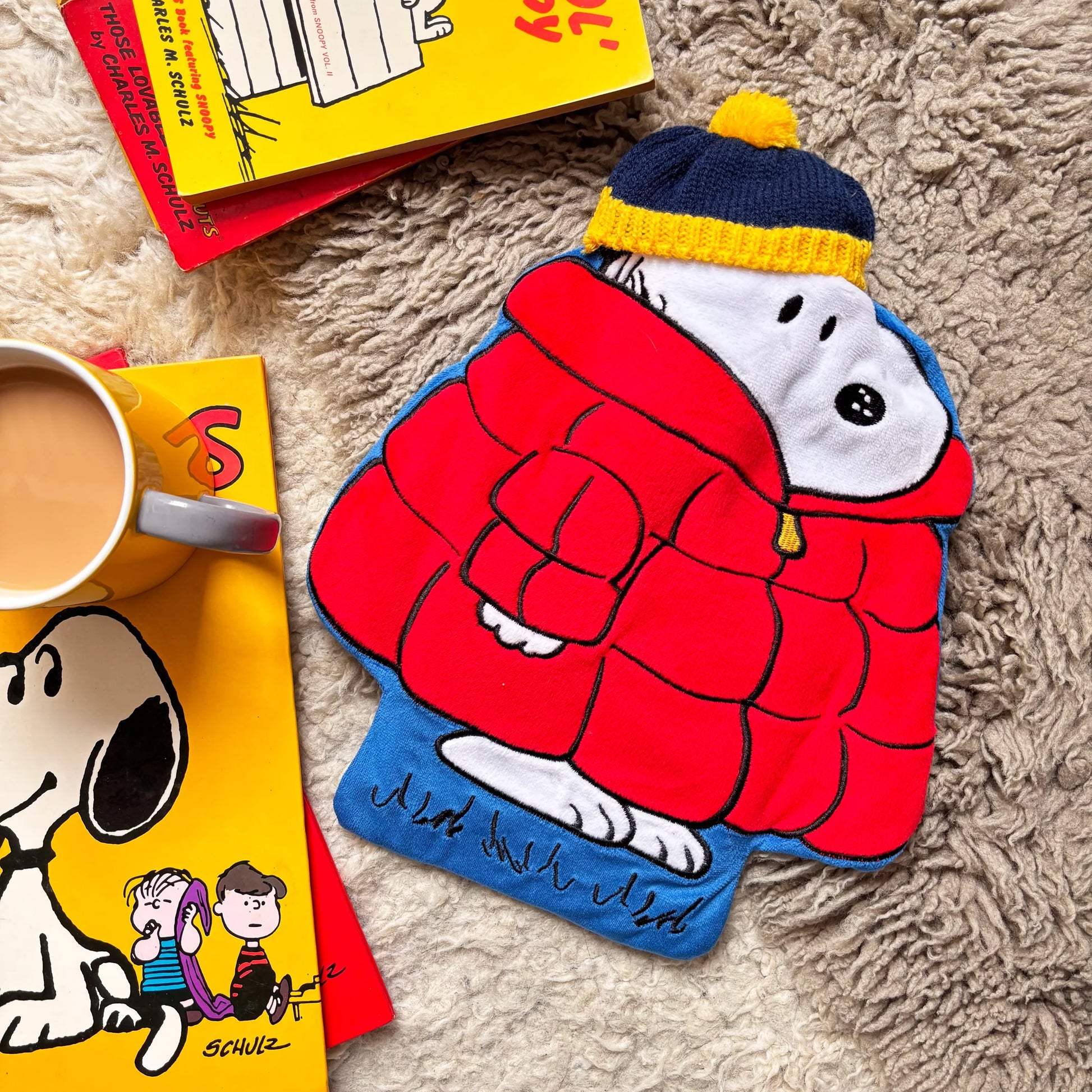 Peanuts Snoopy Puffa Hot Water Bottle on a rug with Peanuts books and mug of tea.