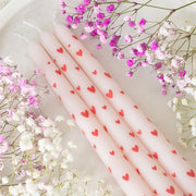 Four pink taper candles with red love hearts surrounded by pink and white gypsophila flowers, styled flat lay for romantic Valentine’s Day décor.