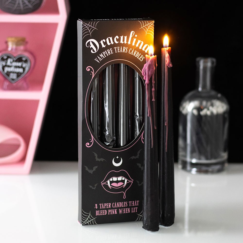 Set of pink-drip vampire taper candles burning next to gothic-themed packaging on a white surface with Halloween decor in background.
