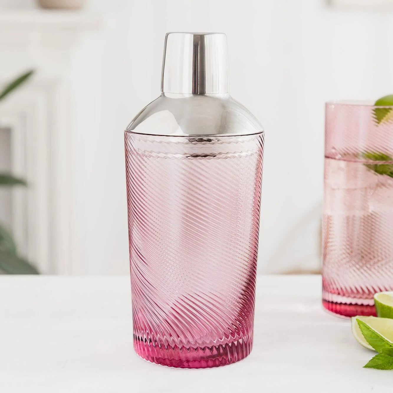 Pink glass cocktail shaker with twisted swirl design and shiny steel top, styled with matching glassware and green leaves.