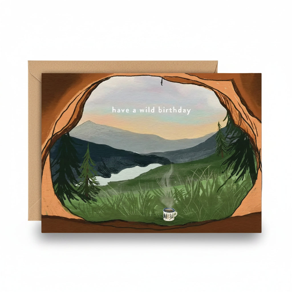 A greeting card with a kraft envelope, featuring an illustrated mountain landscape seen through a cave opening, with the text 'have a wild birthday' and a small mug with a candle at the bottom.