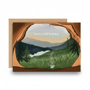 A greeting card with a kraft envelope, featuring an illustrated mountain landscape seen through a cave opening, with the text 'have a wild birthday' and a small mug with a candle at the bottom.