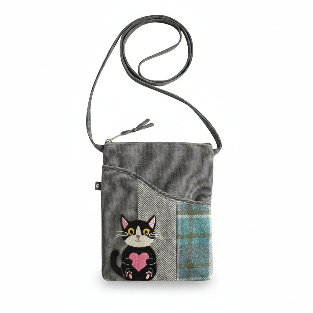 A gray fabric crossbody bag with a black leather strap, featuring a patchwork design with a herringbone pattern, a blue plaid pocket, and an embroidered black-and-white cat holding a pink heart.