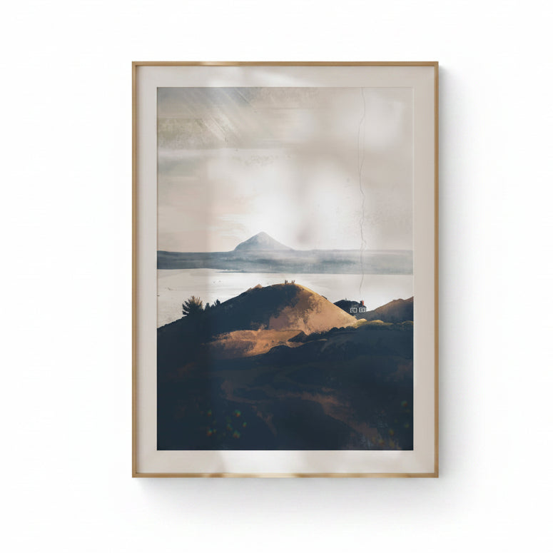 A framed landscape art print featuring a misty mountain range over calm water, with a dark hill in the foreground, displayed on a beige wall with soft leaf shadows.