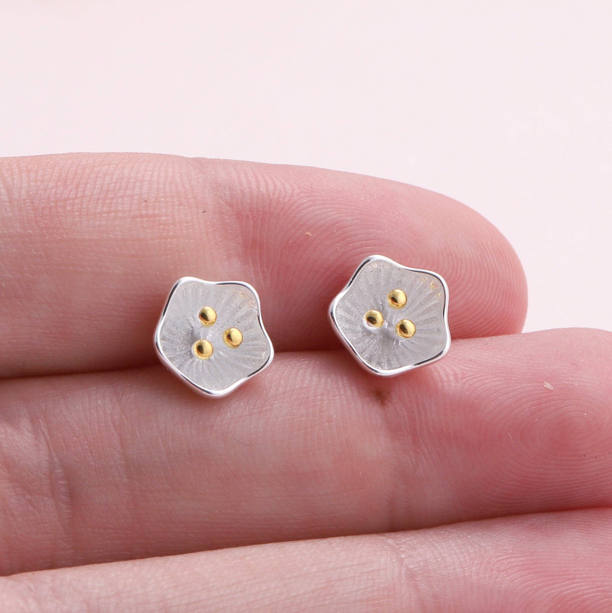 Two silver flower studs with gold bead centres held on fingertips against pink background