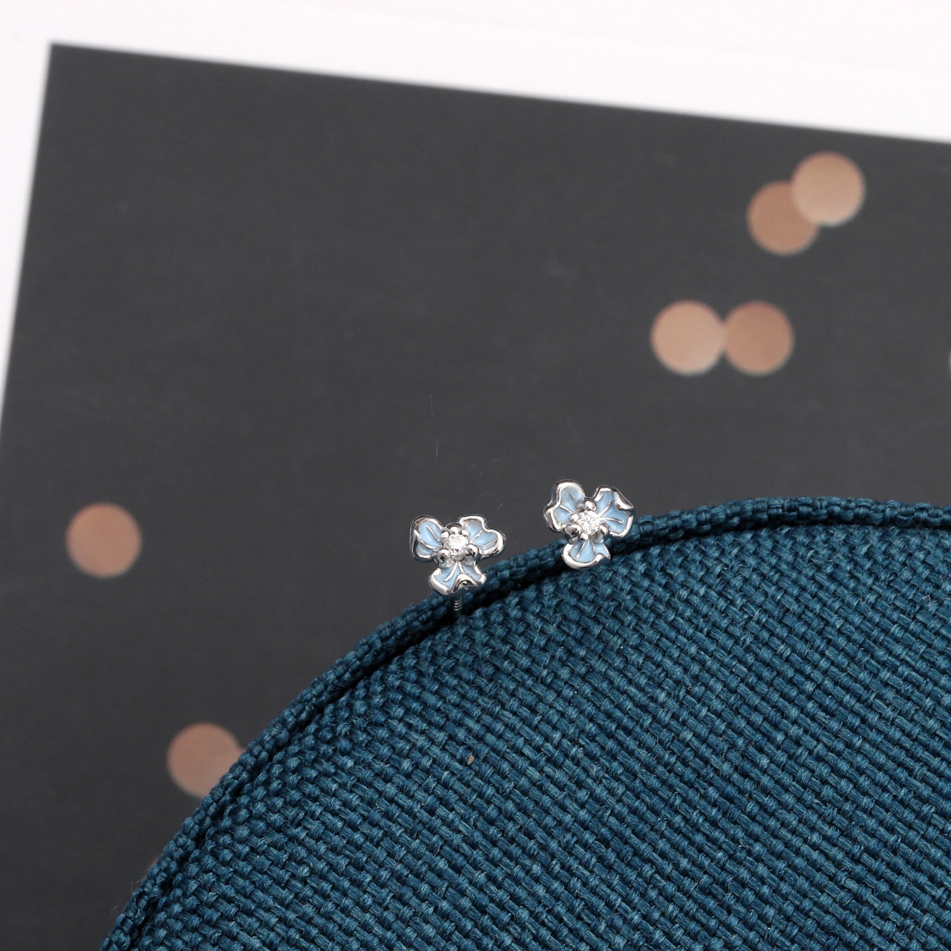 Two tiny blue three-petal flower stud earrings on dark teal fabric with gold dots