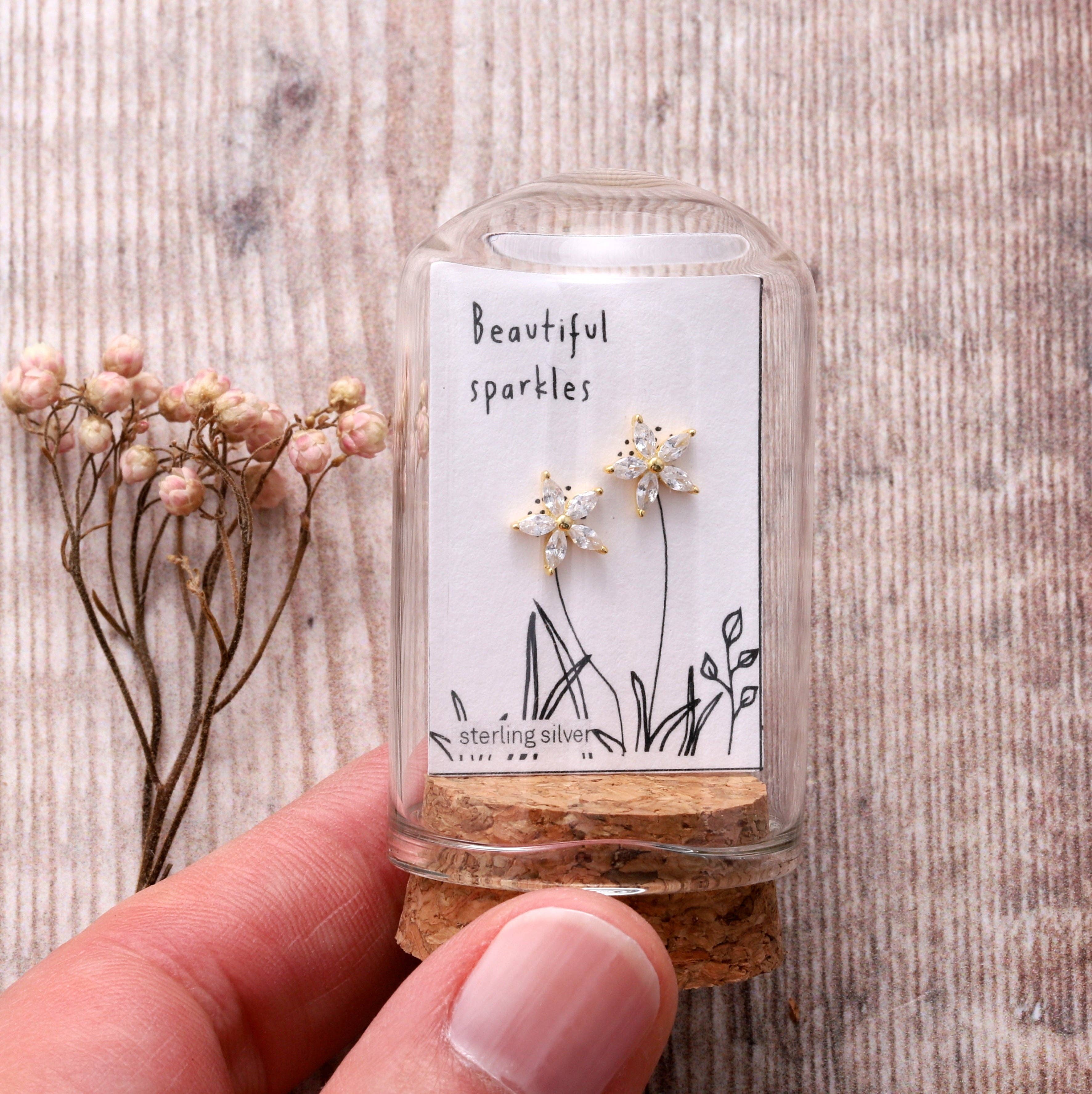 Gold crystal flower stud earrings inside glass terrarium held in hand on grey background