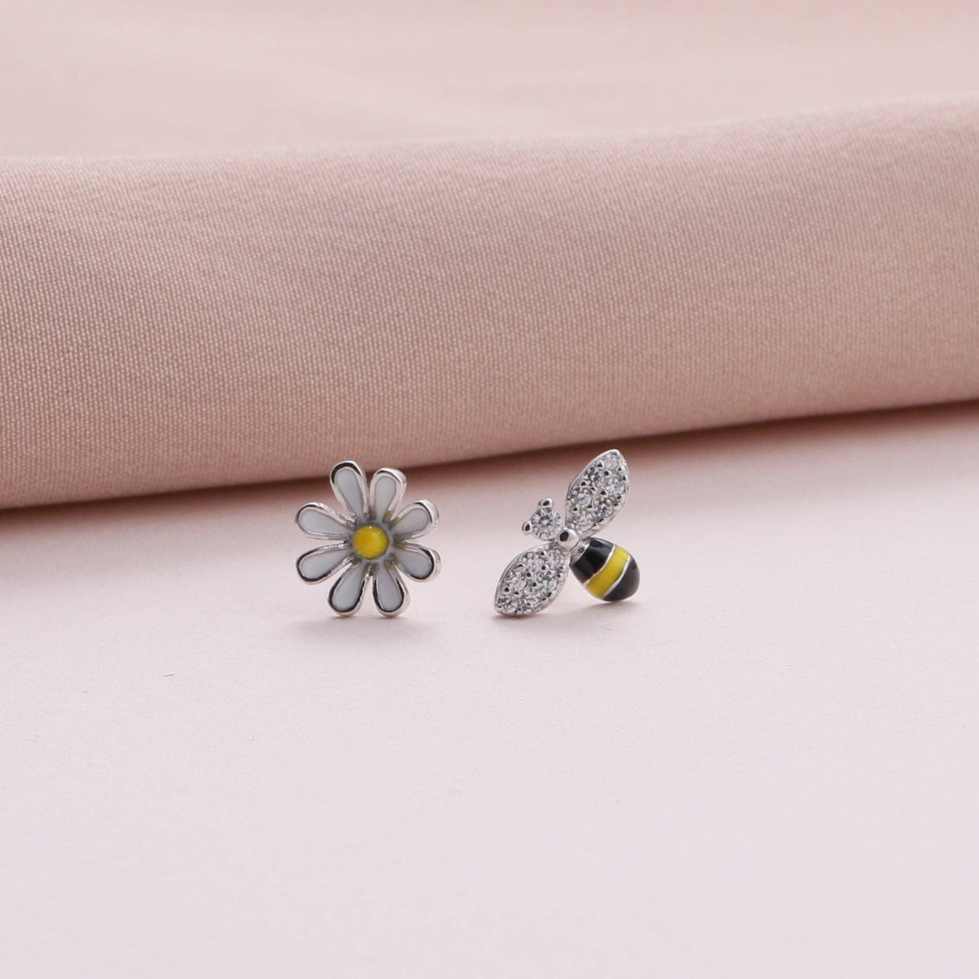 Close-up of enamel bee and daisy stud earrings on dusty pink fabric