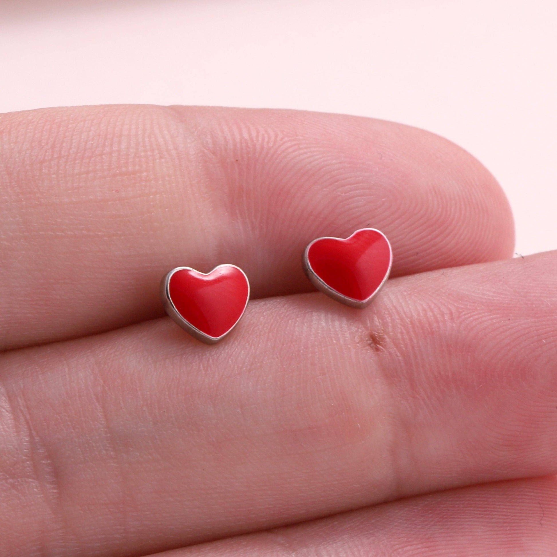 Red enamel heart stud earrings held on fingertips against light background